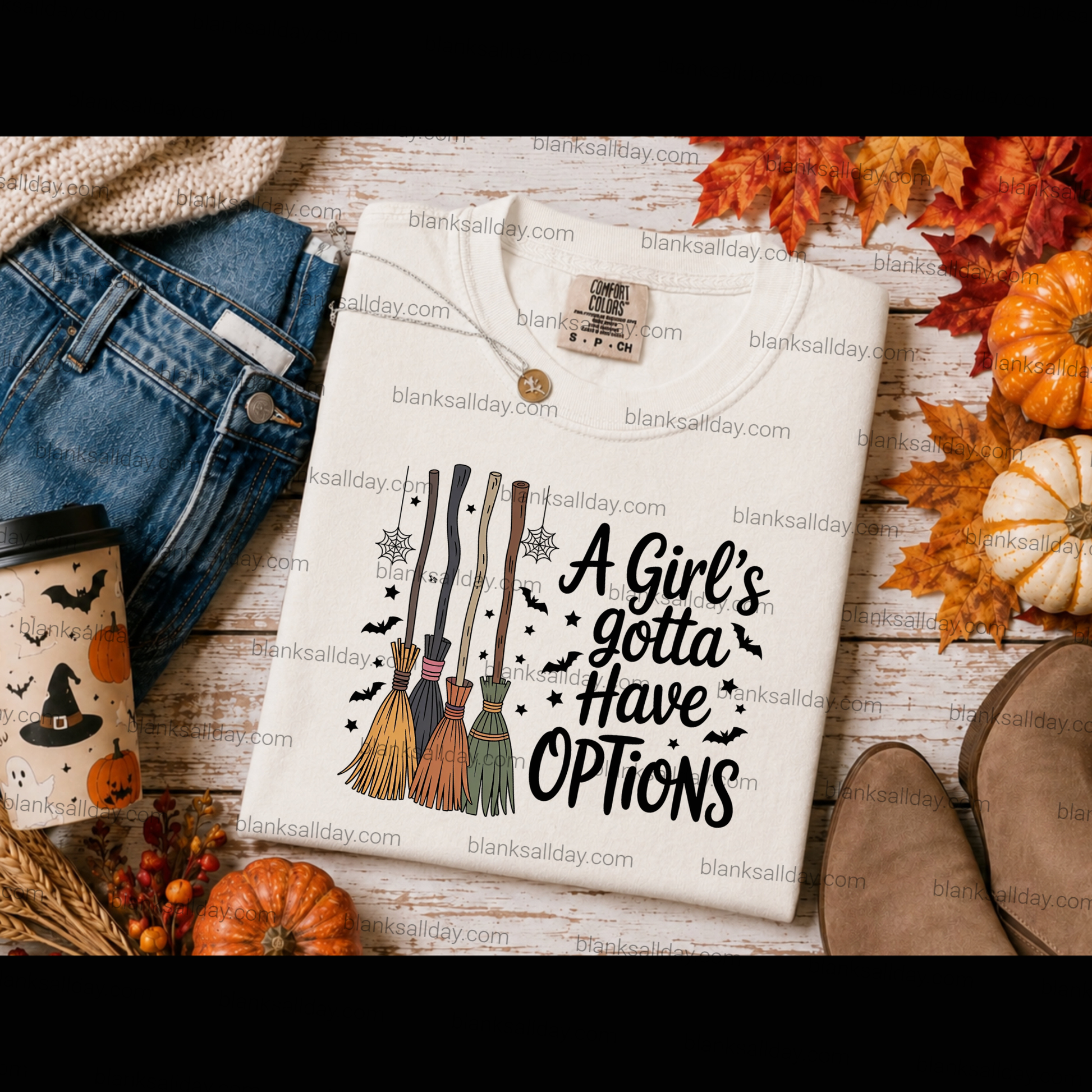 A folded cream graphic T-shirt is styled in a flat lay with autumn props including orange pumpkins, fall leaves, jeans, boots, and a coffee cup. The shirt has a witch-themed graphic with brooms, bats, and the text 'A Girl's gotta Have Options.