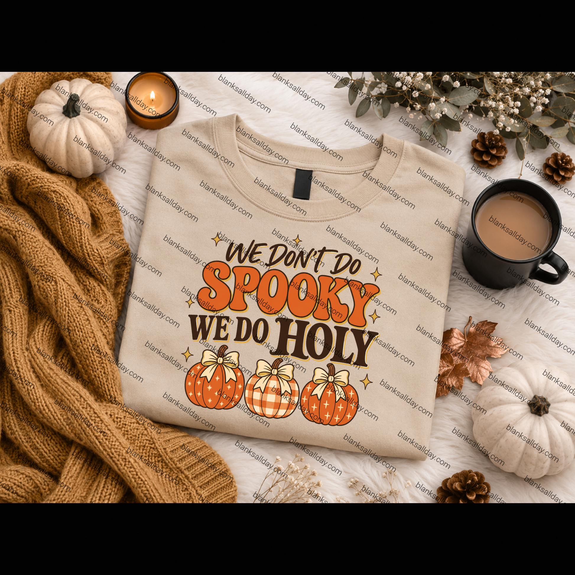 A folded beige graphic T-shirt is styled in a cozy autumn flat lay with a tan knit blanket, small pumpkins, a candle, a mug of coffee, and dried flowers. The shirt reads 'WE DON'T DO SPOOKY WE DO HOLY' in orange and brown lettering above three orange pumpkins.