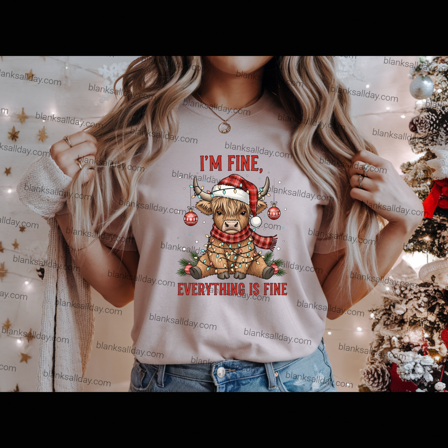 A woman wearing a light beige graphic t-shirt with a Christmas design that reads 'I'M FINE, EVERYTHING IS FINE' with a cartoon sheep in a Santa hat. The background is decorated with warm string lights and a Christmas tree.