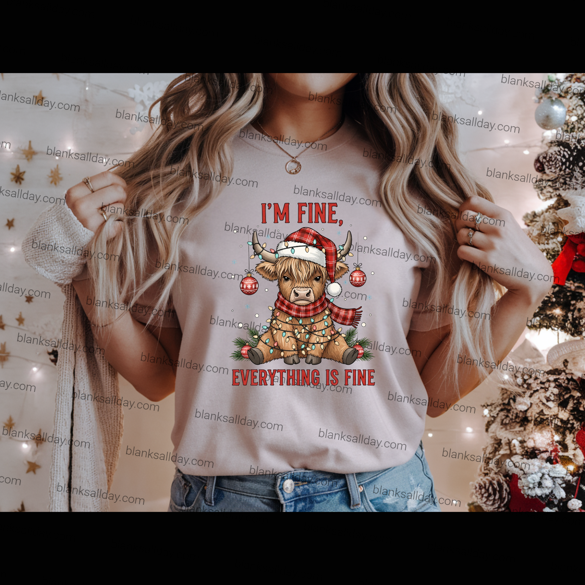 A woman wearing a light beige graphic t-shirt with a Christmas design that reads 'I'M FINE, EVERYTHING IS FINE' with a cartoon sheep in a Santa hat. The background is decorated with warm string lights and a Christmas tree.