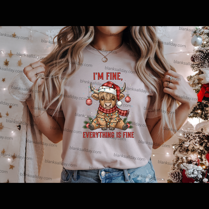 A woman wearing a light beige graphic t-shirt with a Christmas design that reads 'I'M FINE, EVERYTHING IS FINE' with a cartoon sheep in a Santa hat. The background is decorated with warm string lights and a Christmas tree.