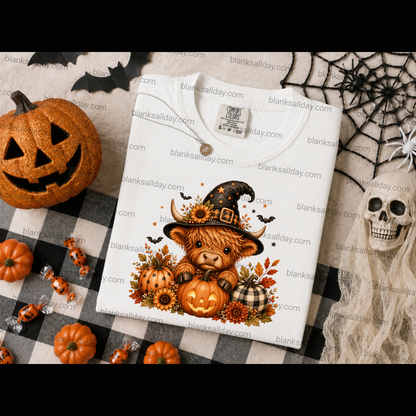 A folded white T-shirt with a Halloween-themed cartoon cow graphic sits on a black-and-white checkered blanket surrounded by pumpkins, spiderwebs, a skull, and candy. The shirt graphic shows a cow in a witch hat holding jack-o'-lanterns with autumn leaves and bats.