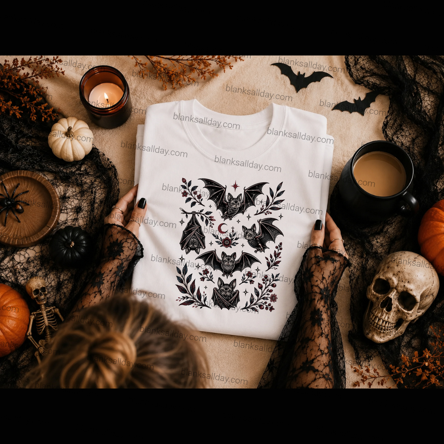 A folded white sweatshirt with a black bat and floral graphic print is held by hands wearing black lace sleeves, surrounded by Halloween props including pumpkins, a skull, a candle, and a mug on a tan surface.