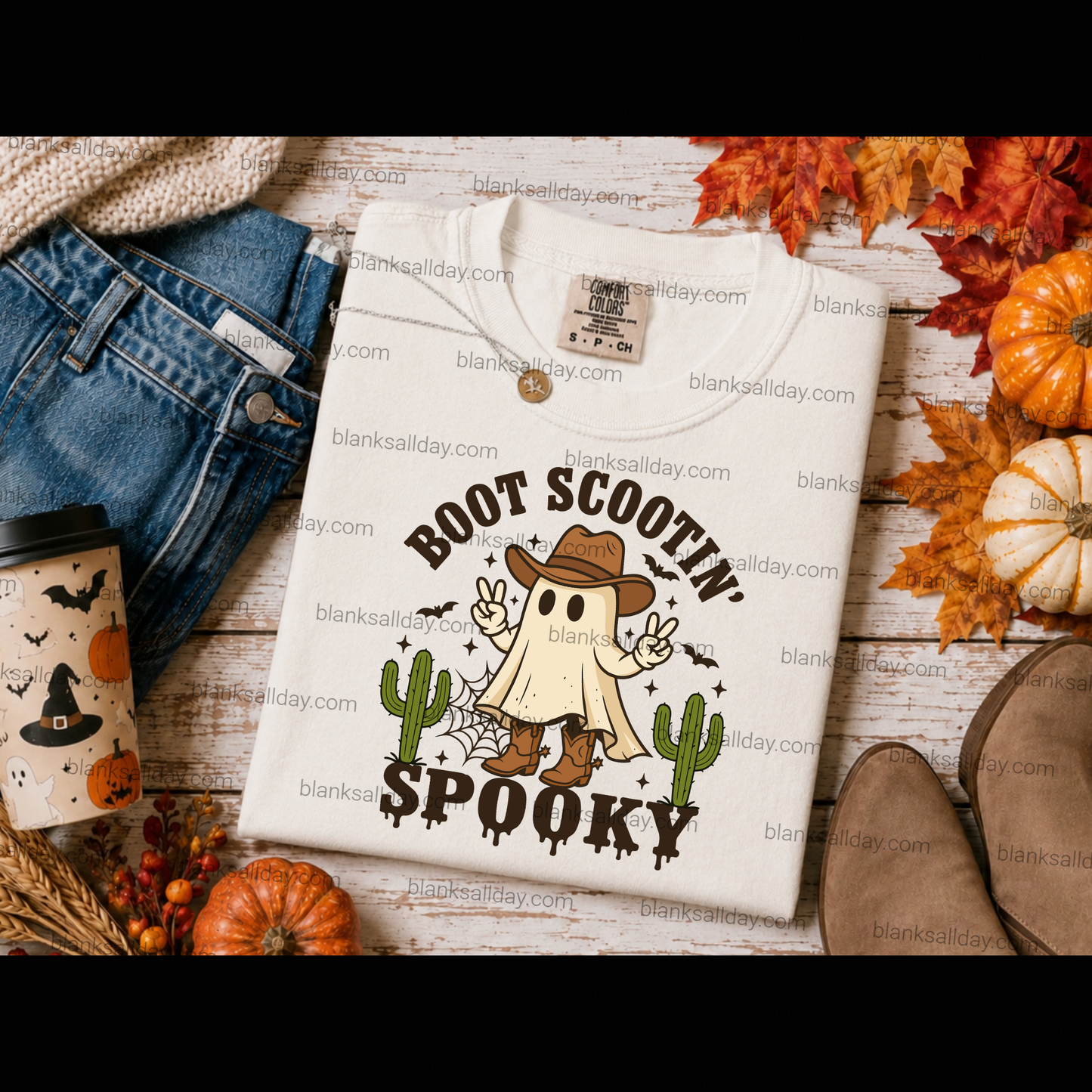 A folded cream-colored t-shirt with a 'Boot Scootin' Spooktacular' Halloween graphic, styled in a flat-lay arrangement with jeans, a coffee cup, autumn leaves, and pumpkins on a rustic wooden background.