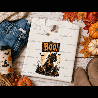 A folded cream-colored t-shirt with a Halloween graphic print featuring a black cat, jack-o'-lanterns, and a 'BOO!' sign, styled in a flat-lay with jeans, a coffee cup, pumpkins, and autumn leaves on a rustic wood background.