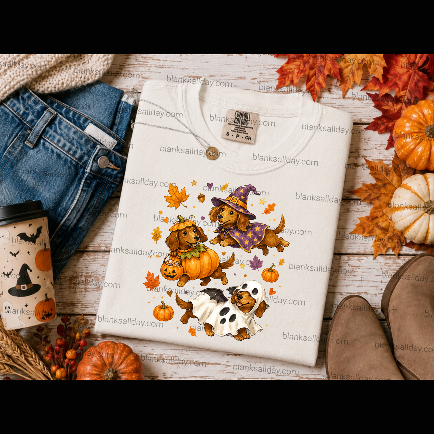 A folded cream-colored t-shirt with a Halloween dachshund graphic, styled in a flat-lay arrangement with jeans, a coffee cup, autumn leaves, and pumpkins on a rustic wooden background.