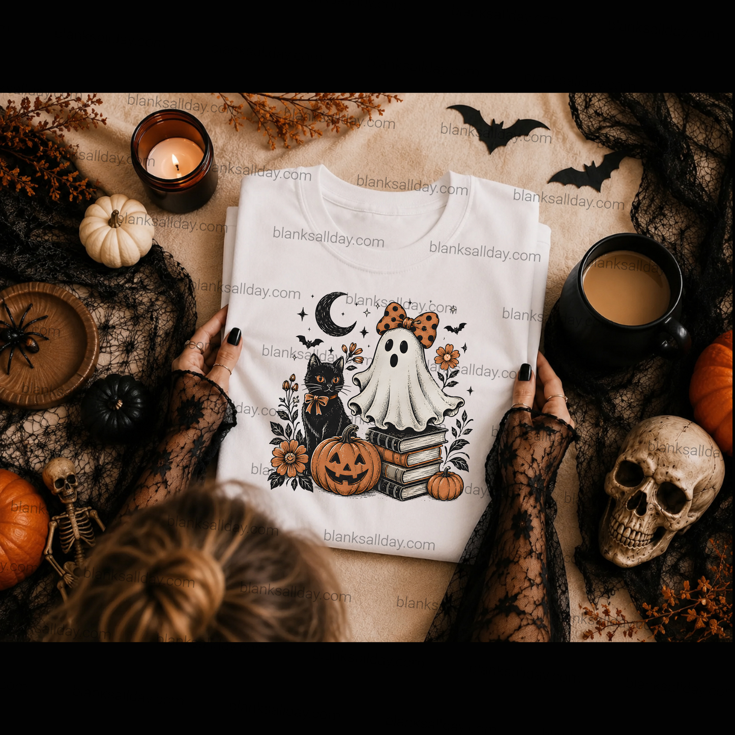 A folded white graphic T-shirt with a Halloween design is held by two hands wearing black lace sleeves, surrounded by autumn and spooky props including a candle, pumpkin, skull, bats, spider decorations, and a mug on a tan fabric background.
