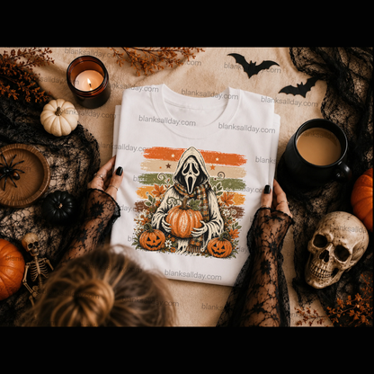 A white graphic t-shirt featuring a hooded skeleton holding pumpkins, styled in a flat lay with hands, a skull, a candle, and autumn decorations on a tan fabric background.