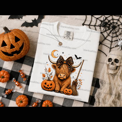 A folded white graphic T-shirt displayed on a black-and-white checkered blanket surrounded by Halloween props, including a carved pumpkin, spiderwebs, a skull, candy, and small pumpkins. The shirt features a cartoon Highland cow wearing a large bow, with pumpkins, a broom, bats, and a crescent moon in an autumn color palette.