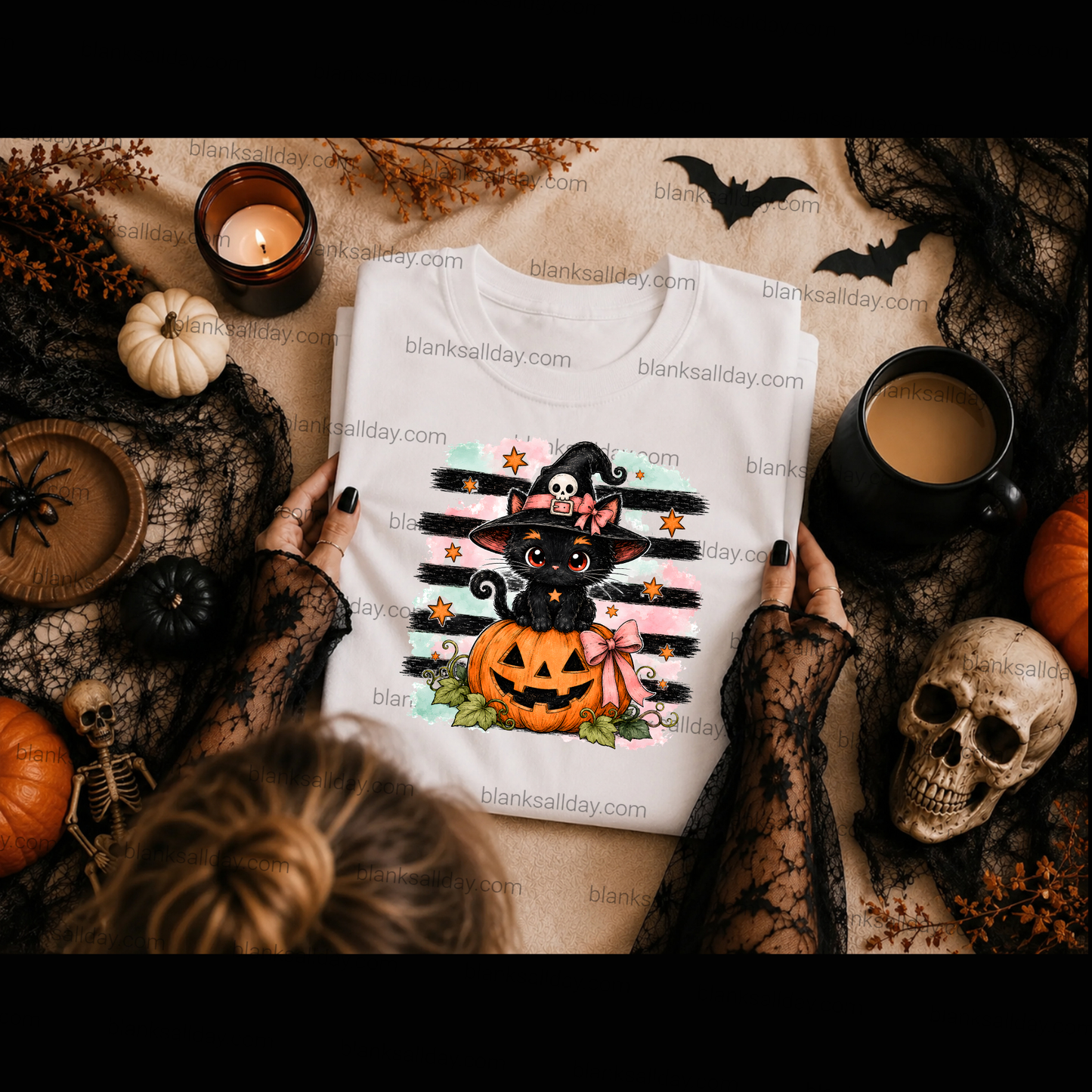 A folded white graphic T-shirt with a Halloween design featuring a black cat in a witch hat above a jack-o'-lantern, held by hands wearing black lace sleeves. The scene is styled as a flat lay with a candle, pumpkin, skull, and other autumn decorations on a tan fabric background.