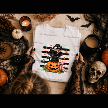 A folded white graphic T-shirt with a Halloween design featuring a black cat in a witch hat above a jack-o'-lantern, held by hands wearing black lace sleeves. The scene is styled as a flat lay with a candle, pumpkin, skull, and other autumn decorations on a tan fabric background.
