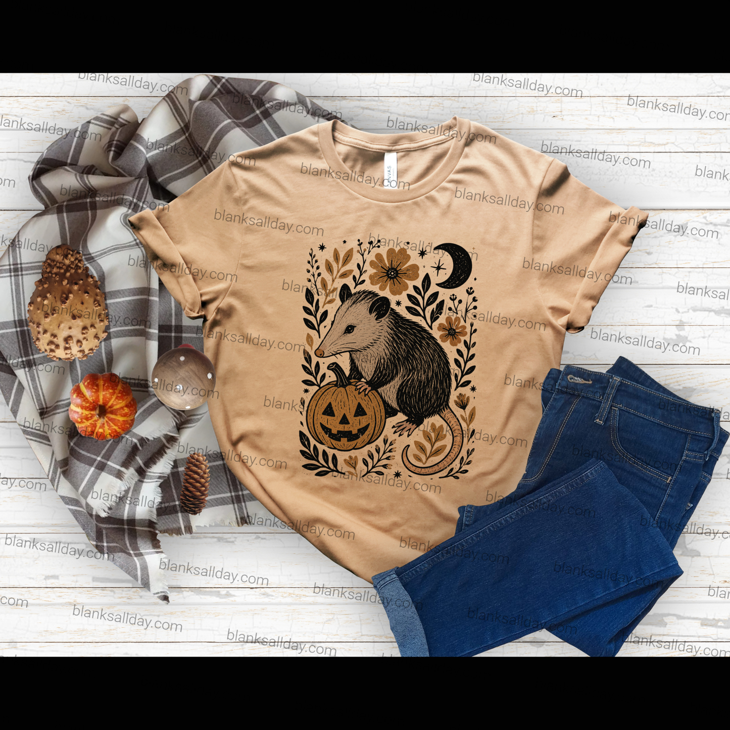 A tan graphic T-shirt laid flat on a white wood surface with a plaid scarf, small pumpkins, pinecones, and blue jeans around it. The shirt has a large fall-themed animal print showing a raccoon beside a jack-o'-lantern with flowers, leaves, and a crescent moon.