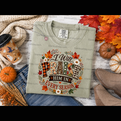 A folded sage green graphic T-shirt is styled in an autumn flat lay with jeans, a coffee cup, boots, pumpkins, and fall leaves. The shirt features a large graphic that reads 'I Will PRAISE HIM IN EVERY SEASON' surrounded by colorful fall leaves, pinecones, and flowers.