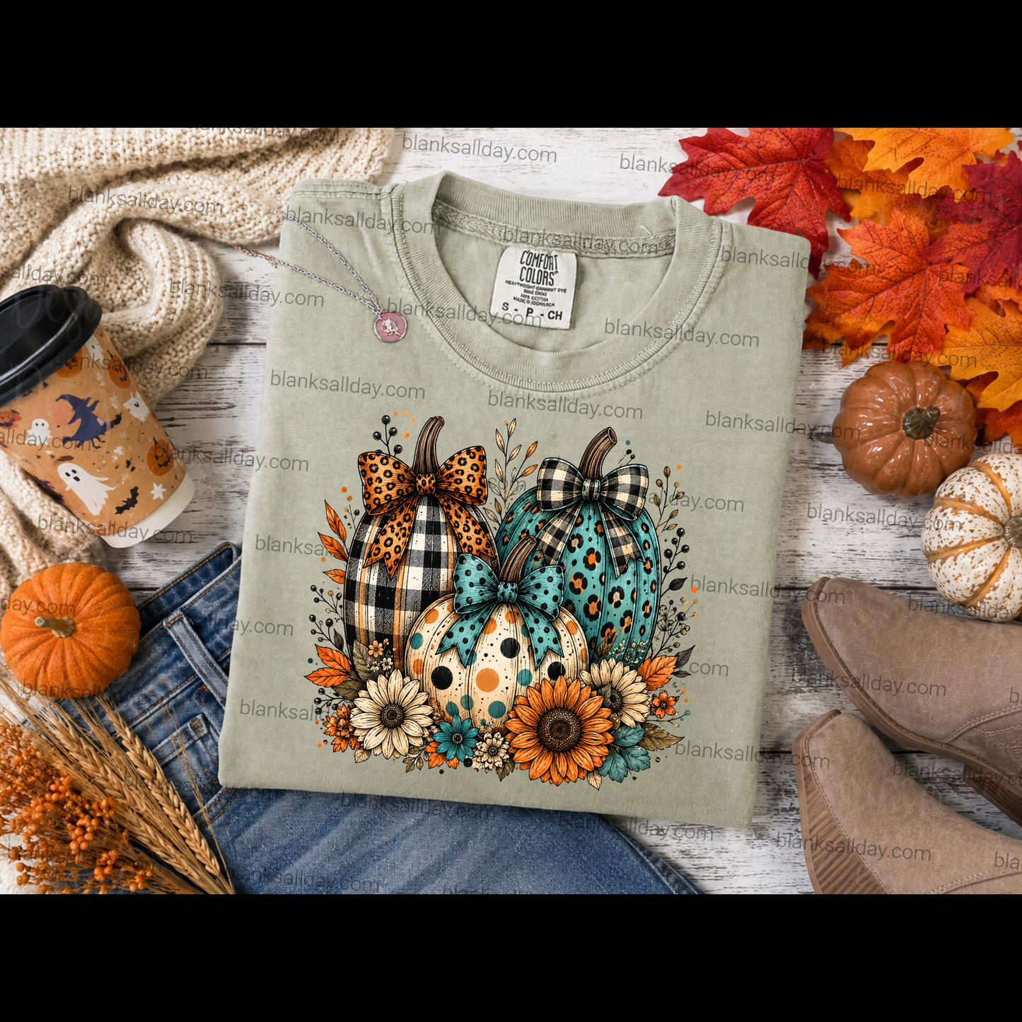 A folded light sage green t-shirt with a colorful autumn pumpkin graphic, styled in a flat-lay with jeans, a coffee cup, fall leaves, and boots.