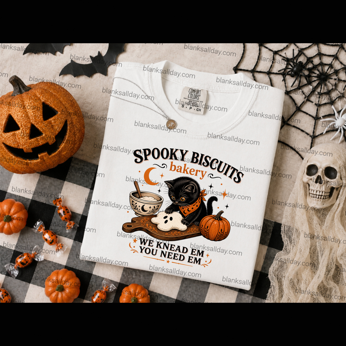 A white short-sleeve T-shirt laid flat on a Halloween-themed backdrop with a pumpkin, spiderwebs, candy, and a skull. The shirt has a printed graphic reading 'Spooky Biscuits bakery' and 'We knead em, you need em' with an illustration of a black cat, pumpkin, cup of coffee, and ghost.